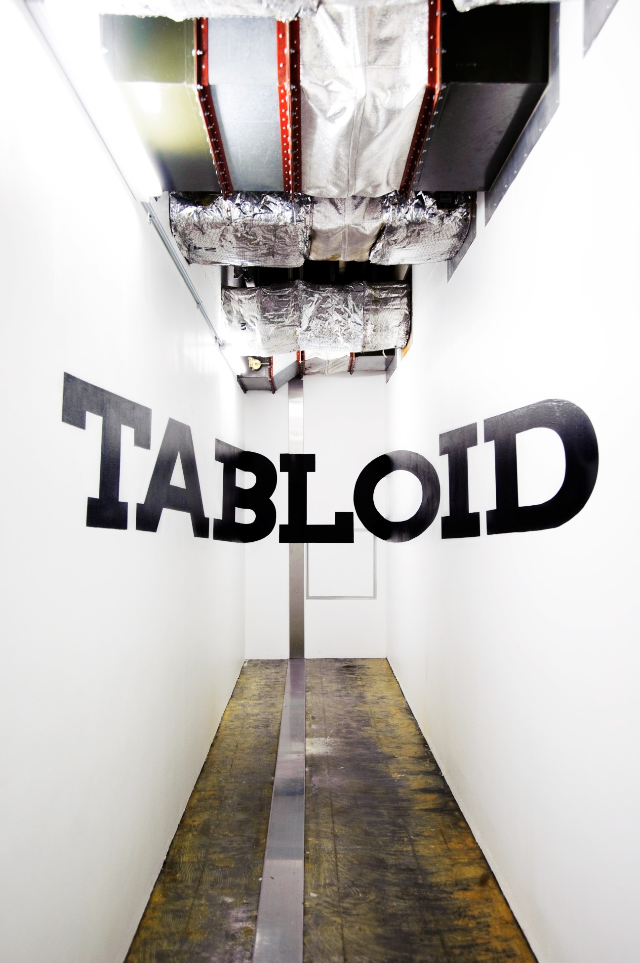Overhead view of Tabloid building entrance with large black lettering on white wall and industrial ceiling elements.