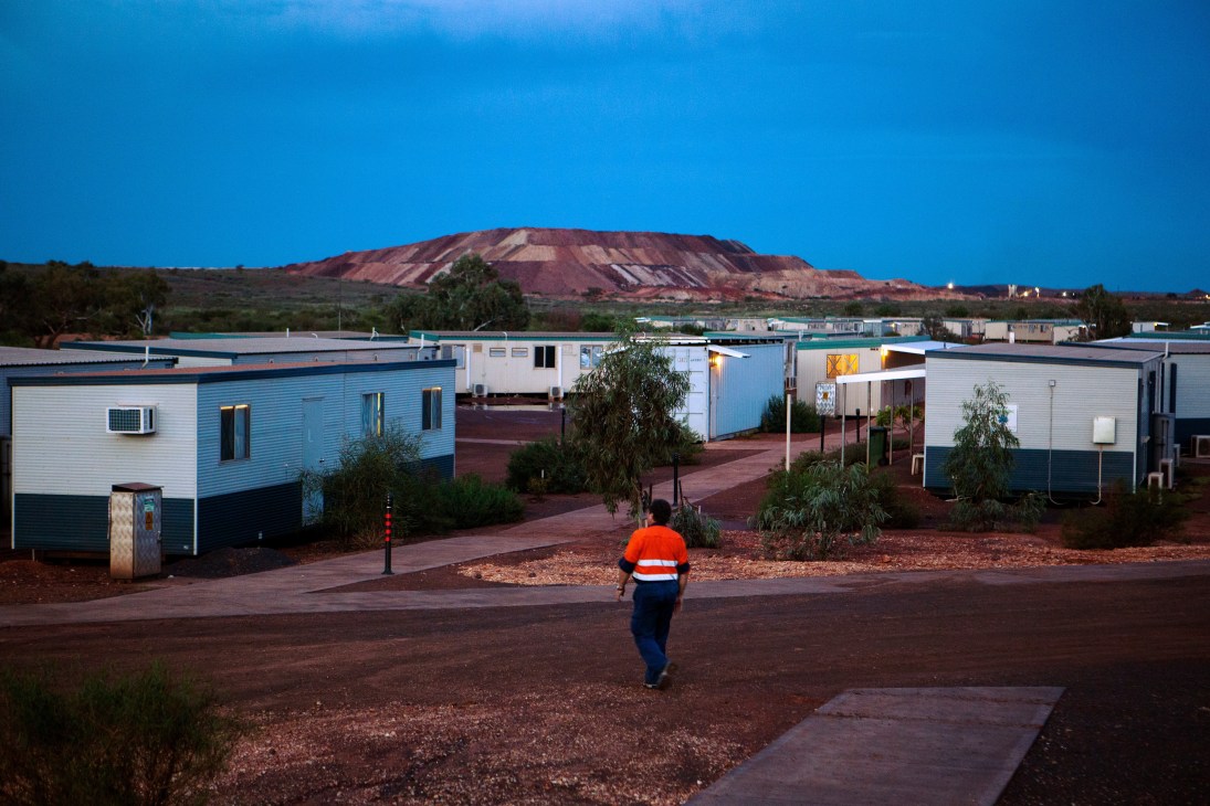 Worker in high-visibility orange vest running through Woodie Woodie mining camp with blue prefabricated dongas and red earth landscape.