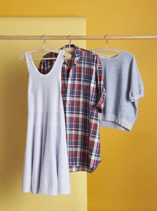 Three garments hanging on wooden hangers against a yellow wall: a striped tank top, a red and blue plaid shirt, and a grey knit sweater.