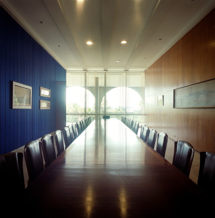 A formal conference room with a long table, black leather chairs, blue and wood-paneled walls, and large windows overlooking gardens.