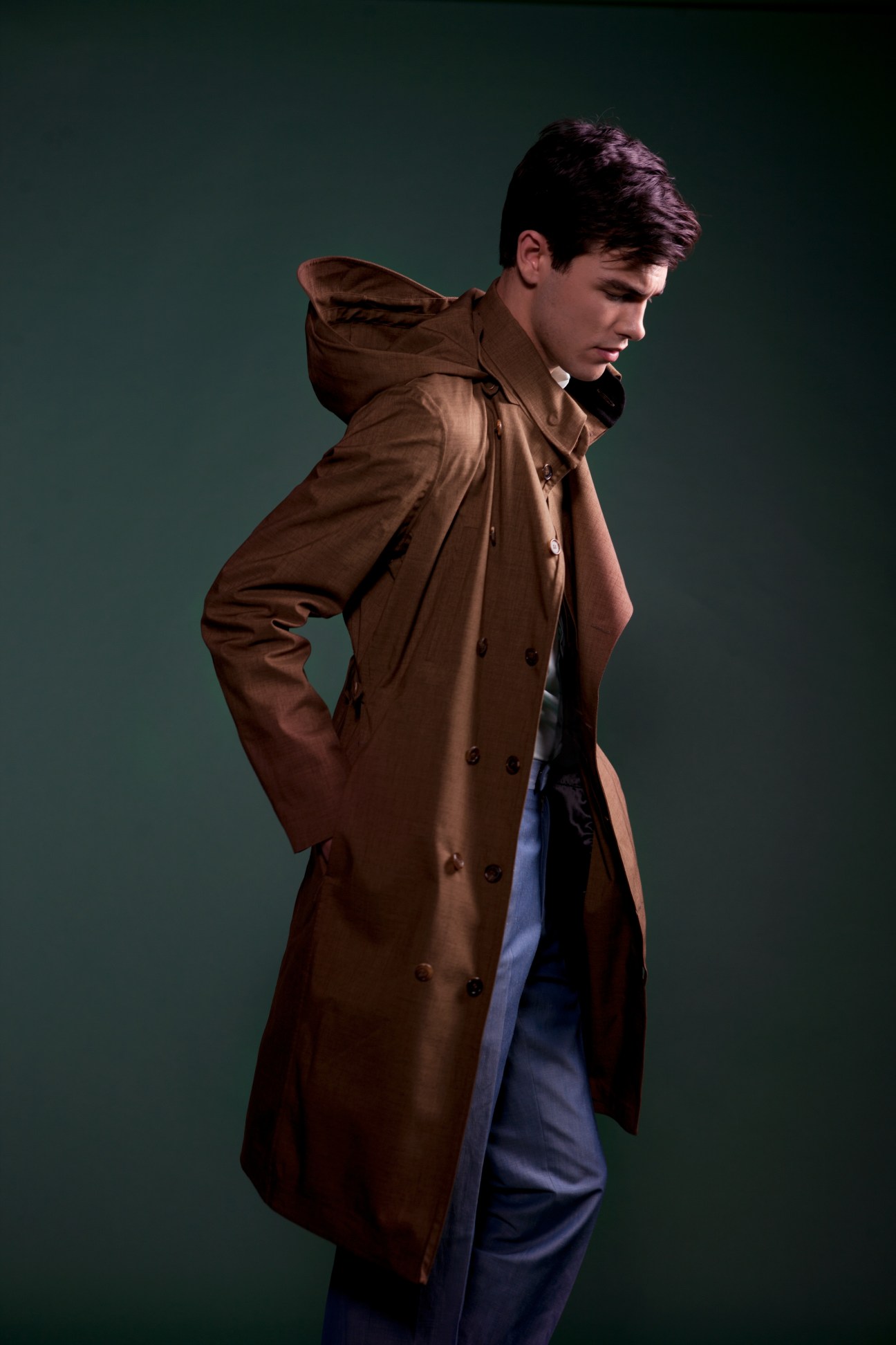 Model wearing a brown double-breasted hooded coat with blue trousers against a teal background.