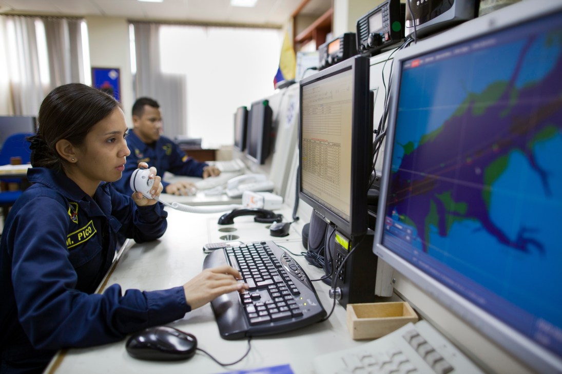 Colombian naval officers monitor radar and radio systems in a control room with multiple computer screens displaying maps.