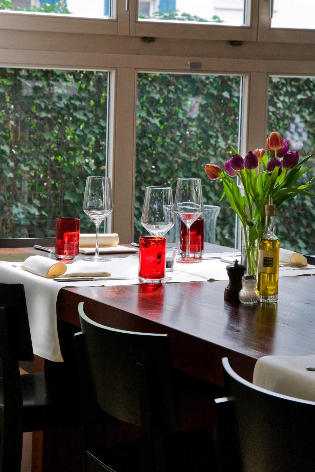 Modern restaurant dining table set with glasses, wine, and purple tulips overlooking green foliage.