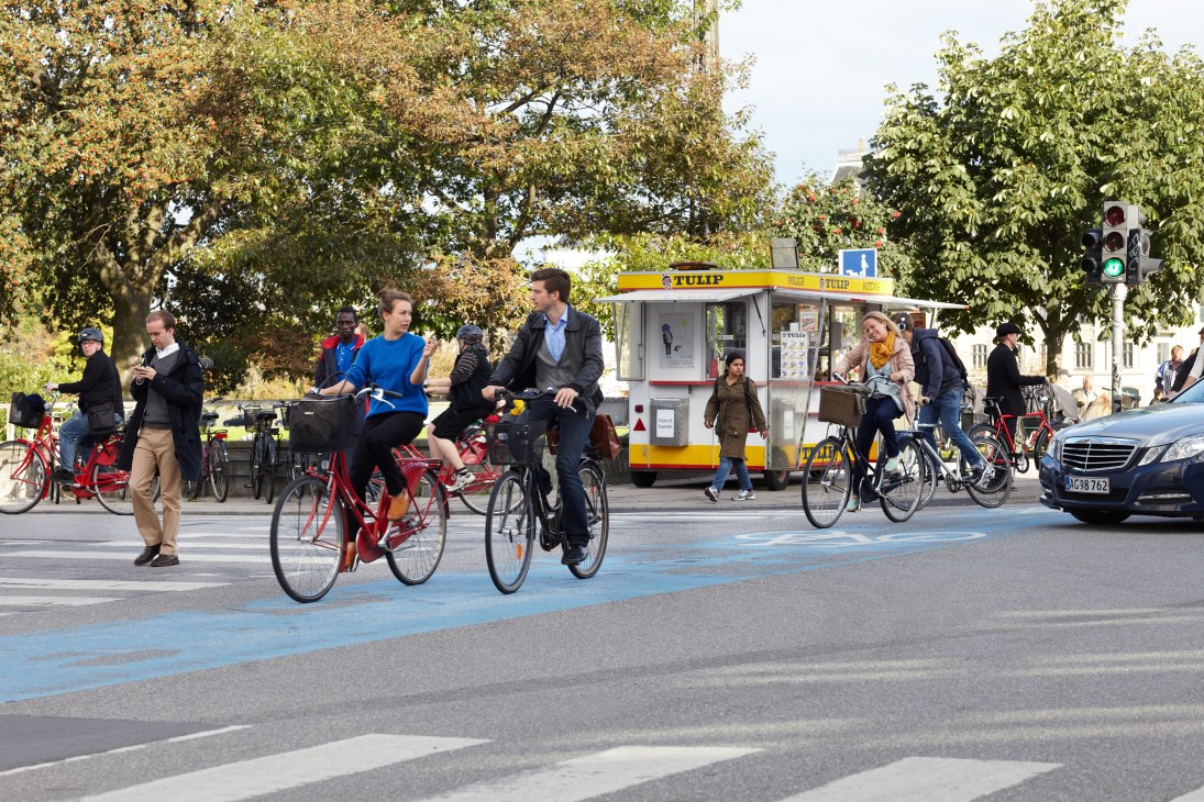 Cyclists ride on a blue bike lane in Copenhagen with a yellow food cart and trees in the background.