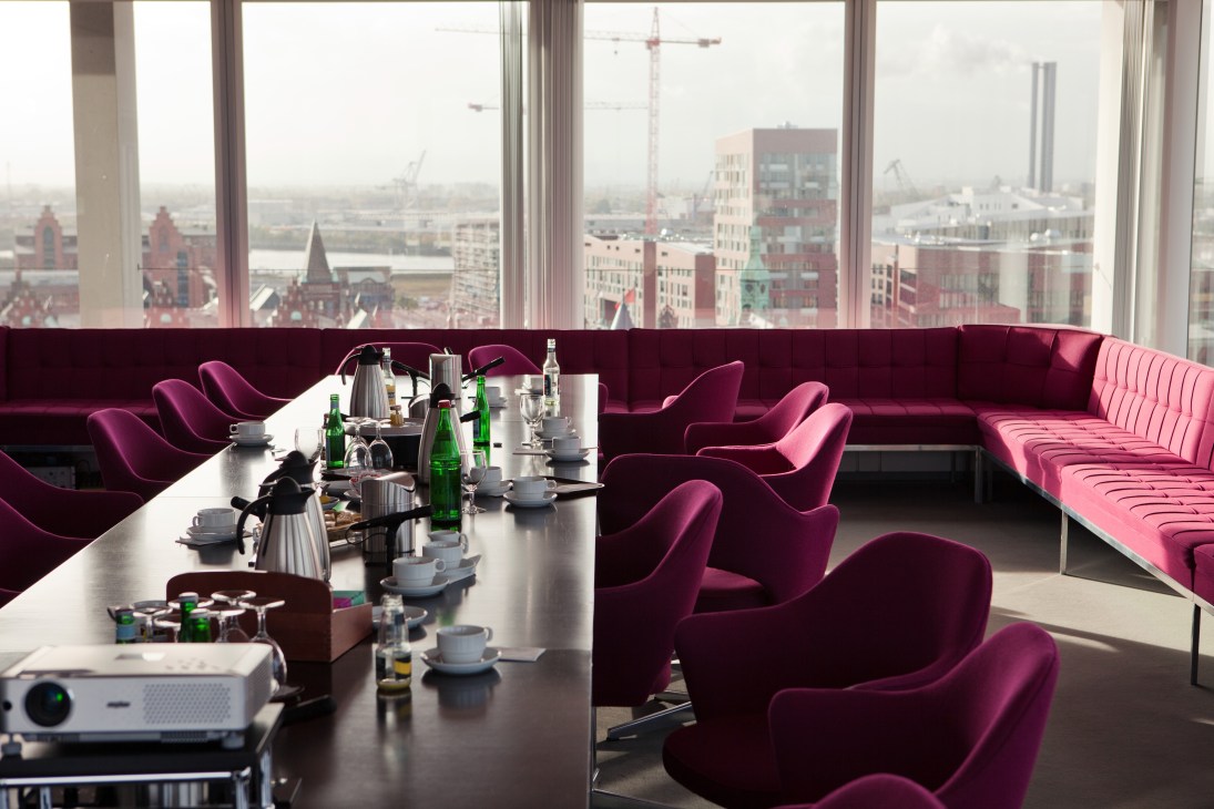 Top-floor conference room with burgundy Verner Panton chairs and sofa, overlooking Hamburg's cityscape through floor-to-ceiling windows.