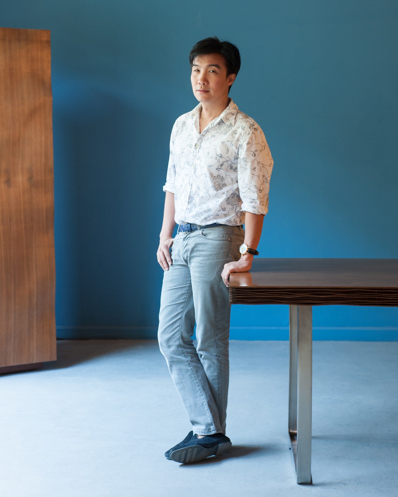 Kenneth Cobonpue stands beside a wooden table against a blue wall in his showroom.