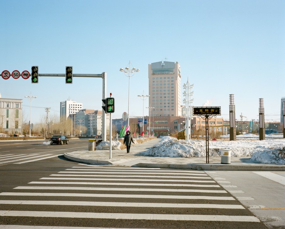 Snowy street intersection in downtown Heihe with traffic lights, pedestrians, and modern apartment buildings under clear blue sky.