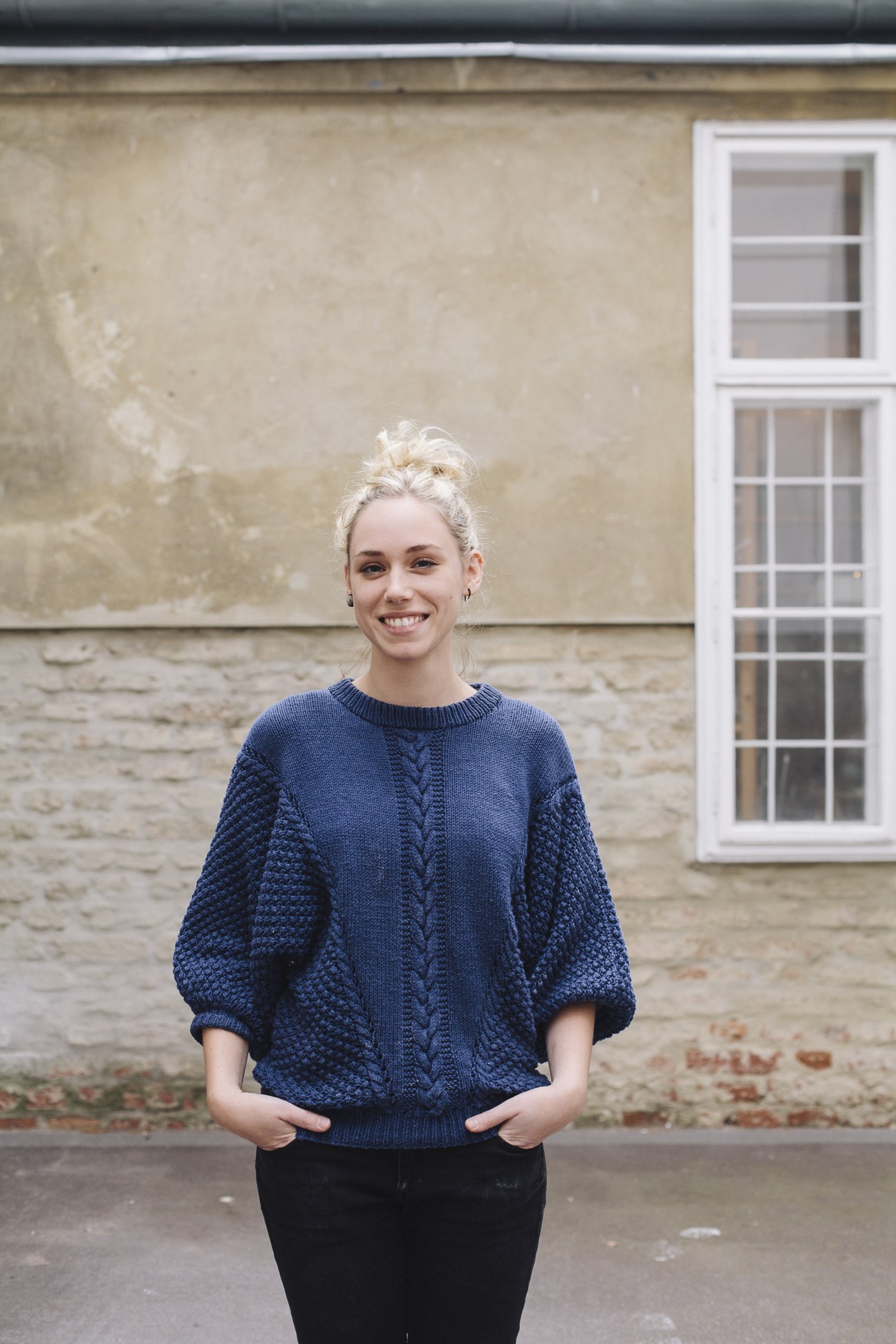 Woman in blue cable-knit sweater smiling at camera outside stone building with white window