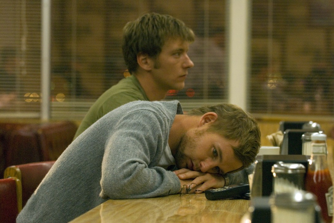 Two men sit at a table in a diner or restaurant, one leaning on the other's shoulder in a casual, intimate moment.