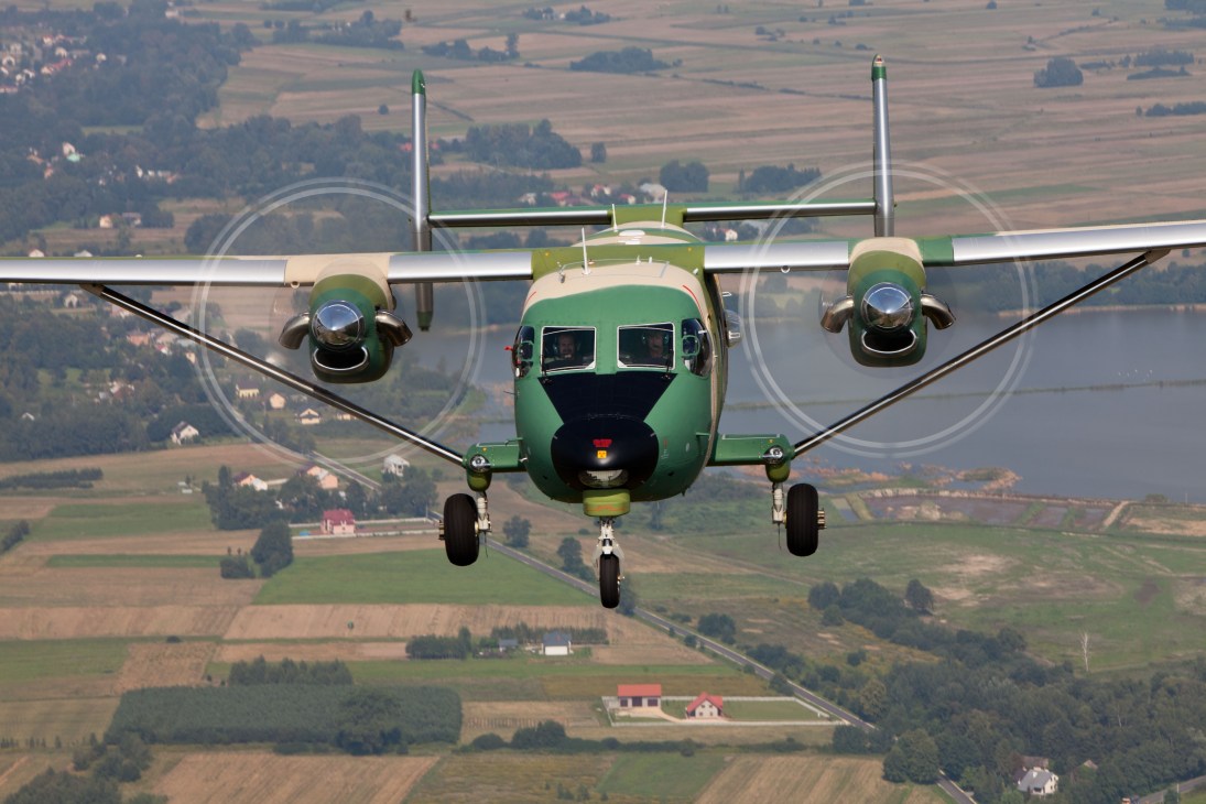 Green and white M28 fixed-wing aircraft flying head-on above rural Polish farmland with forests and villages below.
