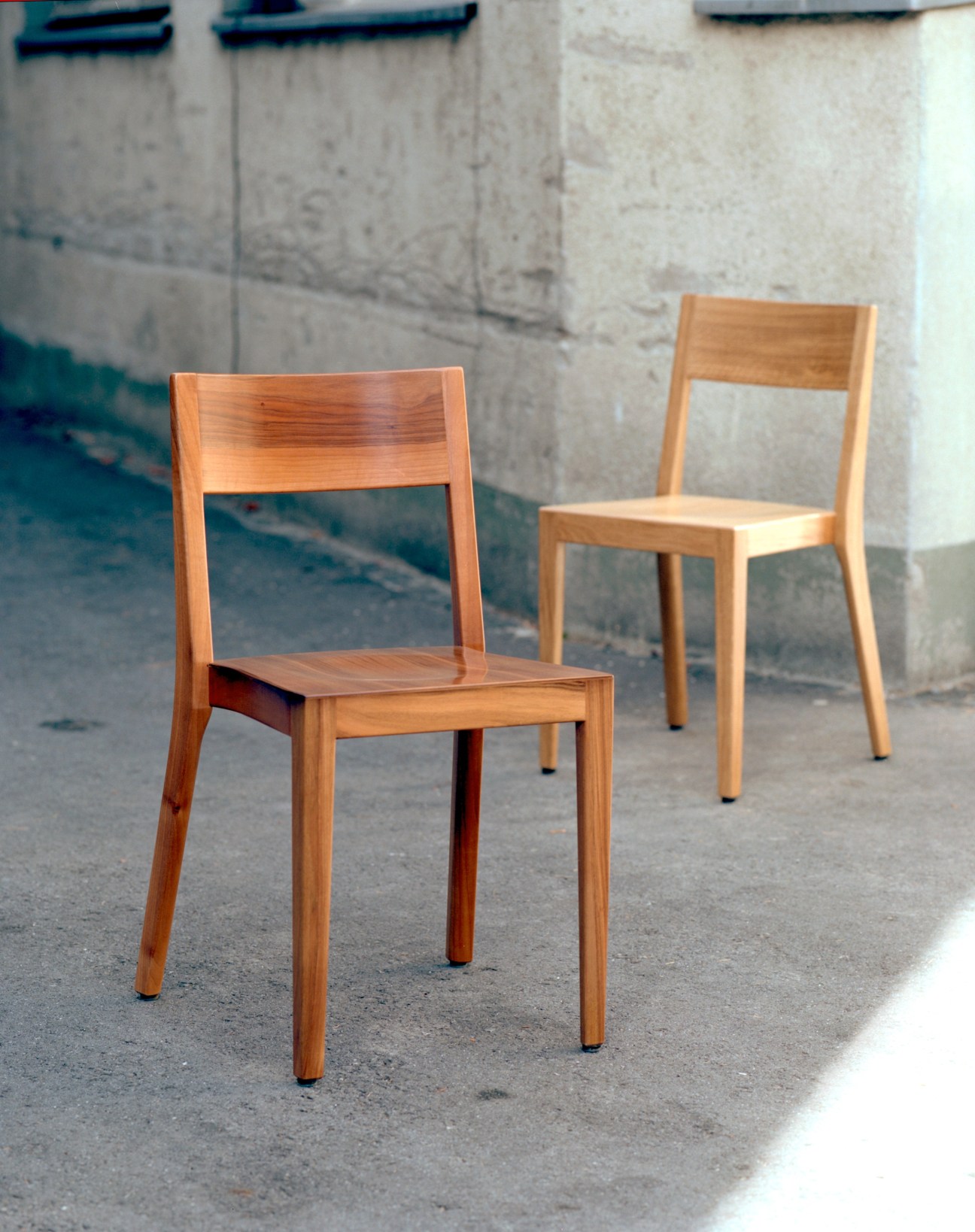 Two wooden Horgenglarus chairs in light wood displayed outdoors against a concrete wall.