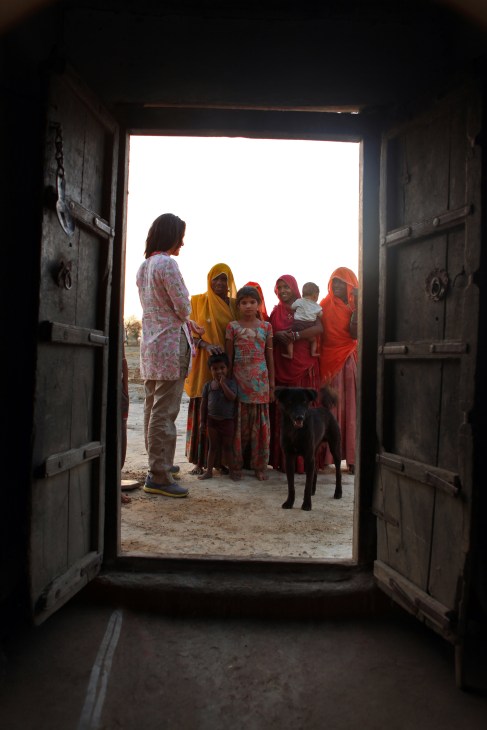Woman in patterned shirt standing in doorway speaking with group of villagers wearing colorful traditional clothing outside in rural Soda, India.