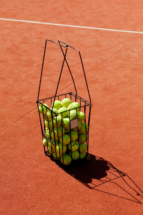 Tennis ball hopper filled with yellow-green tennis balls on a clay court with white line markings.
