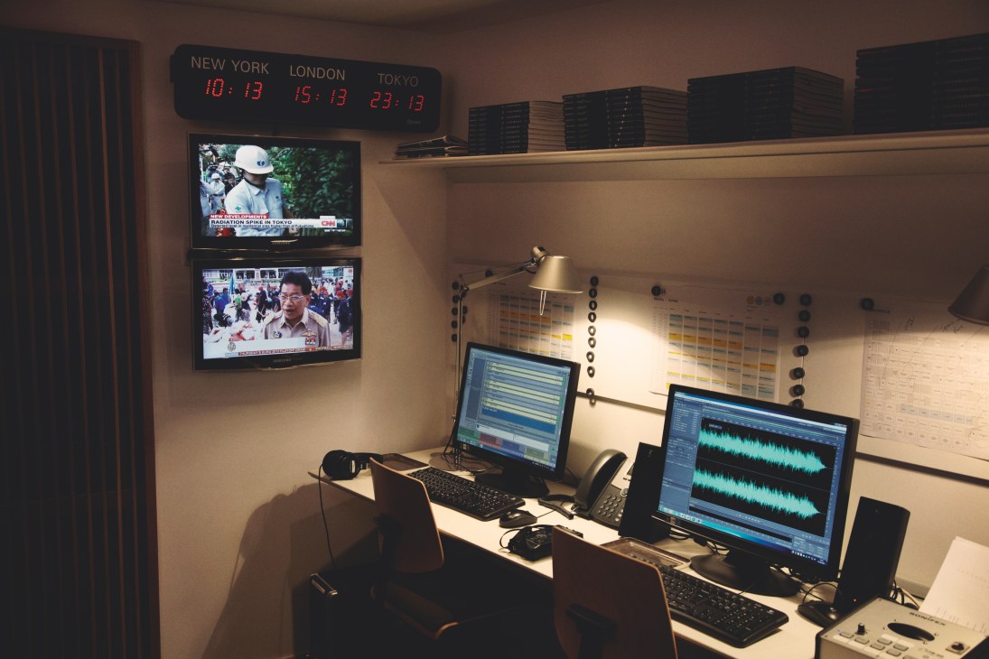Monocle 24 radio studio production area with dual monitors showing audio waveforms and news broadcasts, world clocks above.