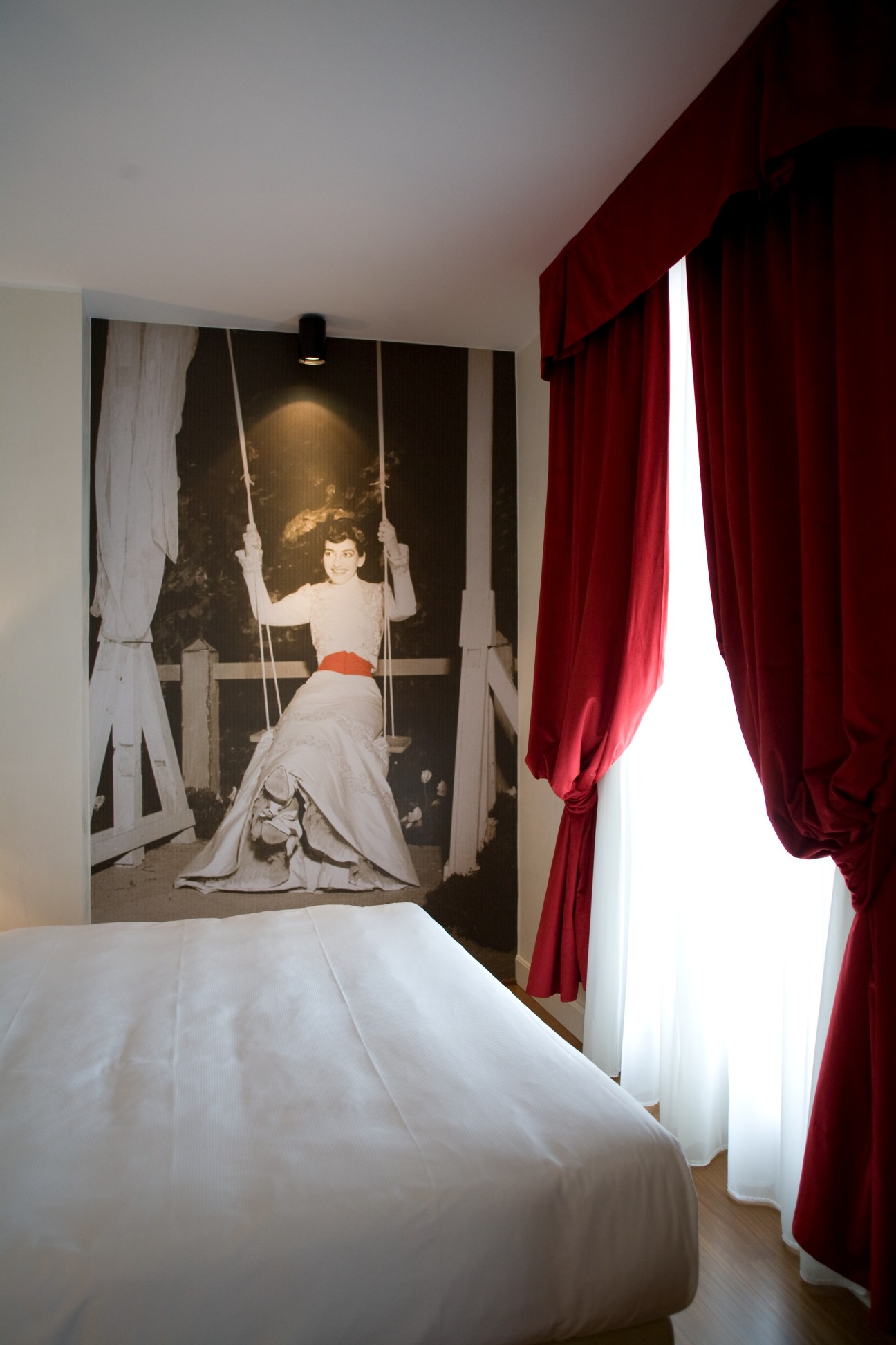 Hotel bedroom with white bed and red curtains facing a large vintage photo mural of a woman on a swing.
