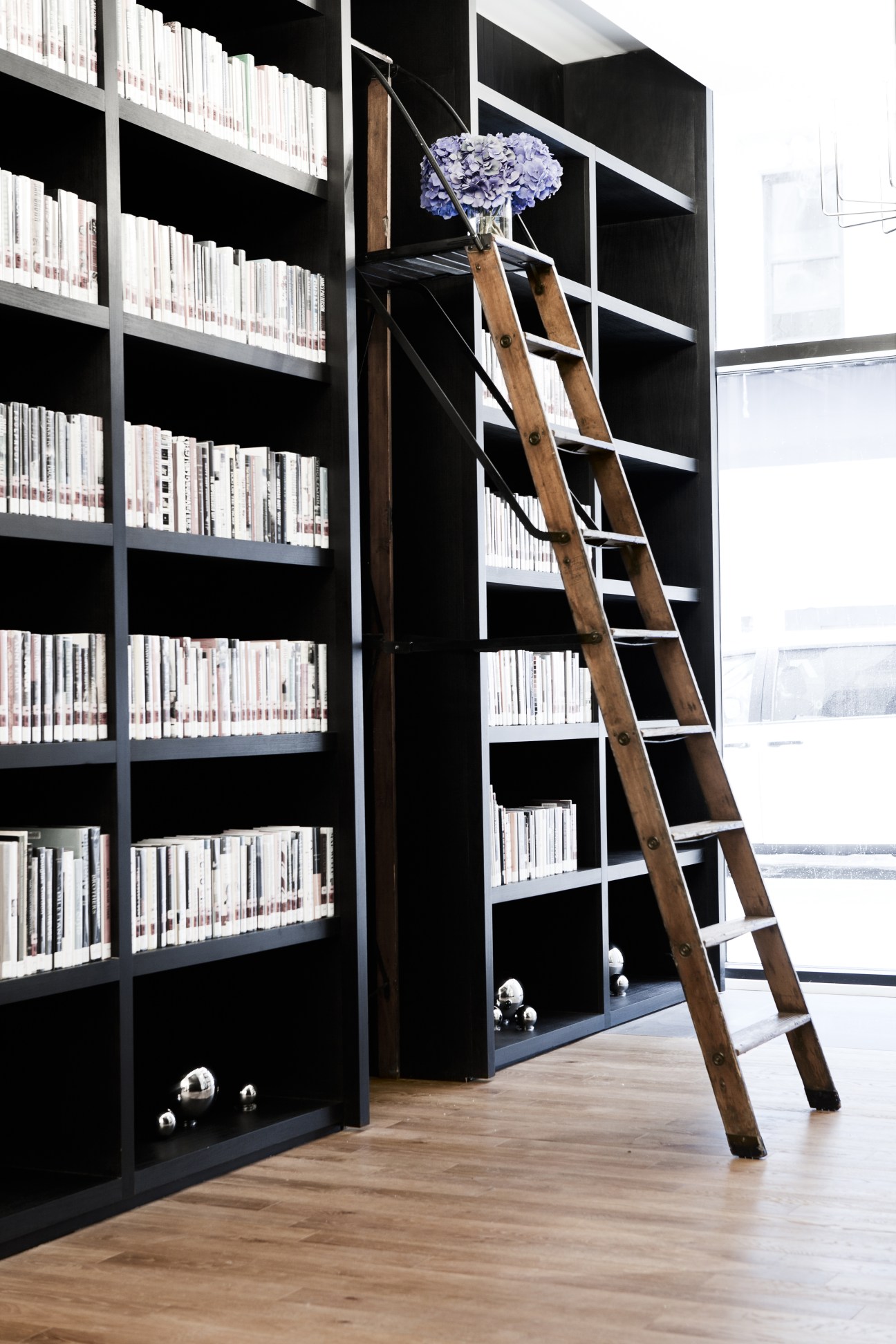 Black shelving unit filled with white books and a wooden library ladder with purple flowers in a vase.