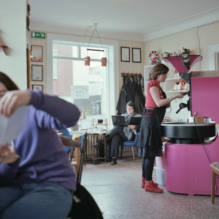 Interior of Kaffi Midja coffee shop with woman at pink counter and customers inside.