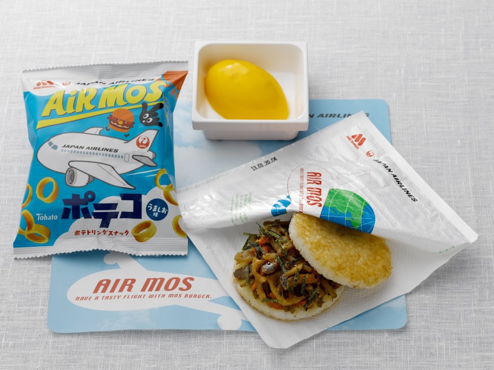 JAL airline meal service including a burger, snack packet, yellow lemon, and branded items on white surface.
