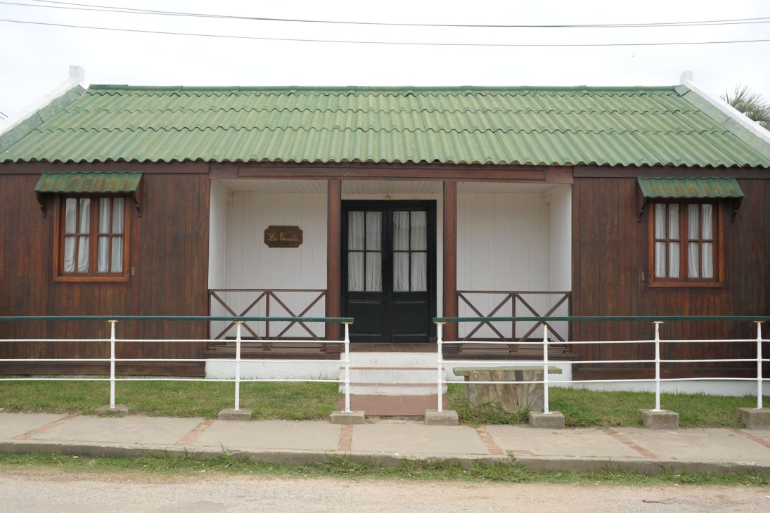 Traditional single-story house with brown wood exterior, green corrugated metal roof, white porch railings and windows in La Pedrera.