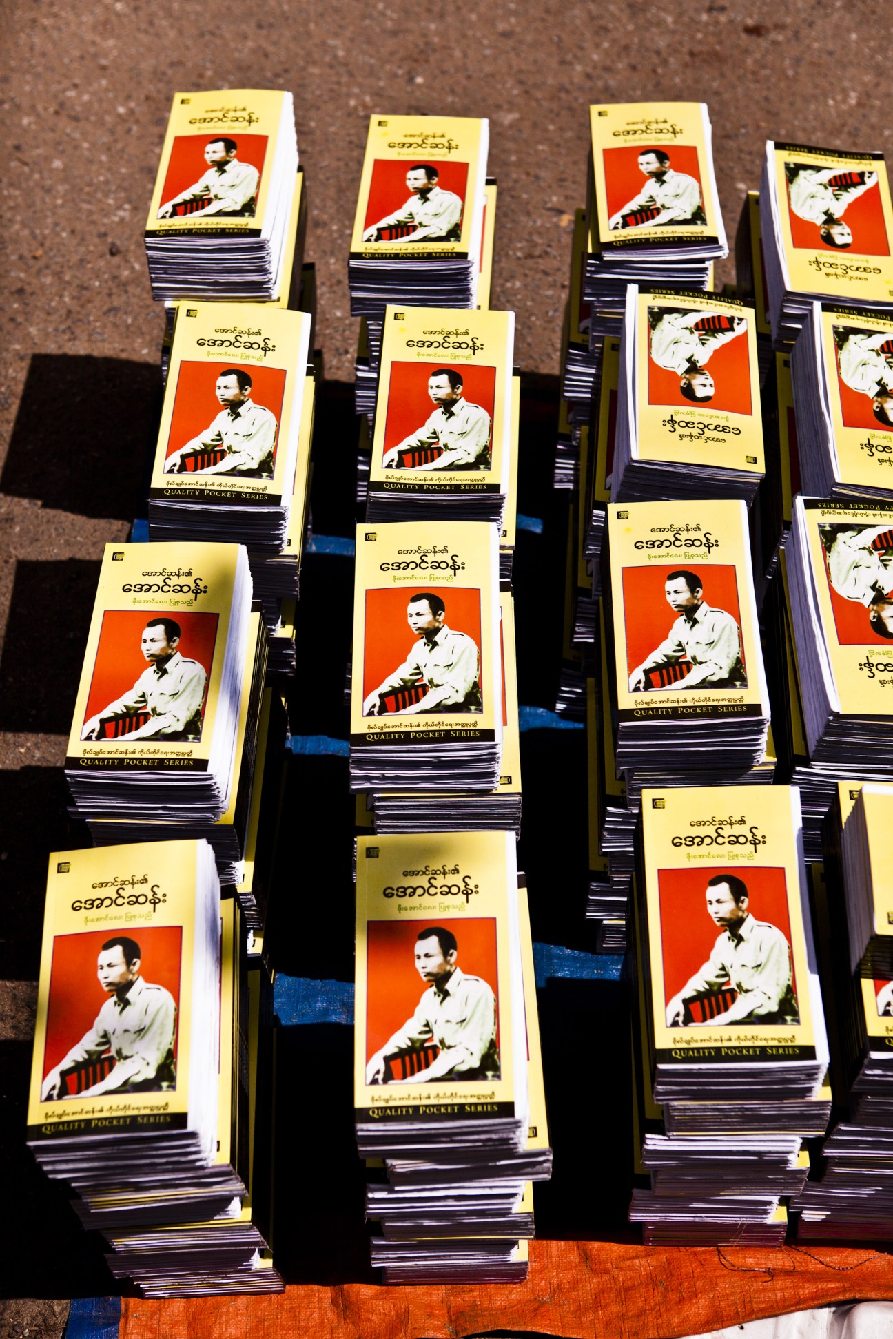Stacks of newly printed books featuring General Aung San on cream and red covers displayed at a market stall.