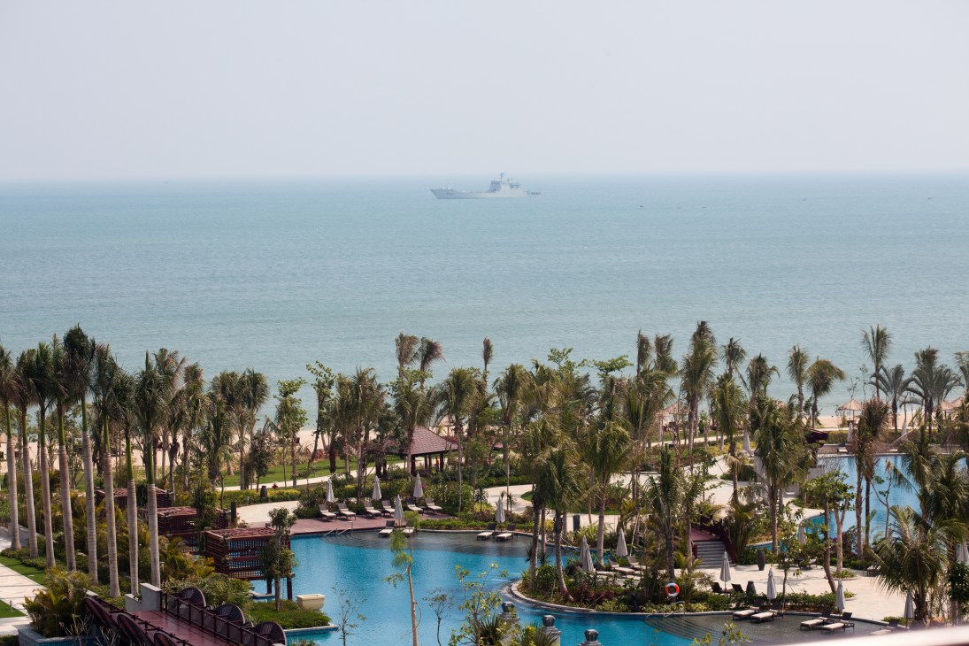 Renaissance hotel gardens with palm trees, pools, and beach overlooking the South China Sea with a military vessel visible offshore.