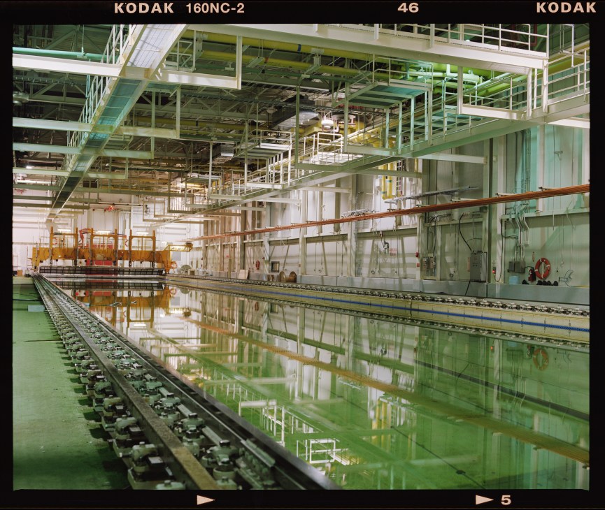 Interior of a large industrial testing tank facility with green-painted infrastructure, overhead cranes, and water tanks for ocean technology research.