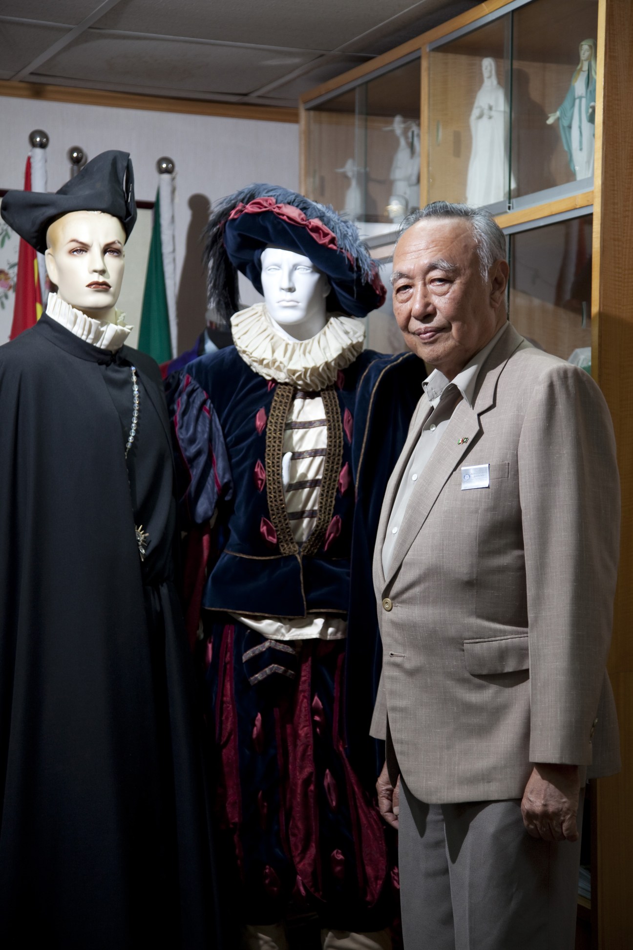 Gary Ngai stands beside two mannequins dressed in elaborate historical costumes with period headpieces in a museum display.