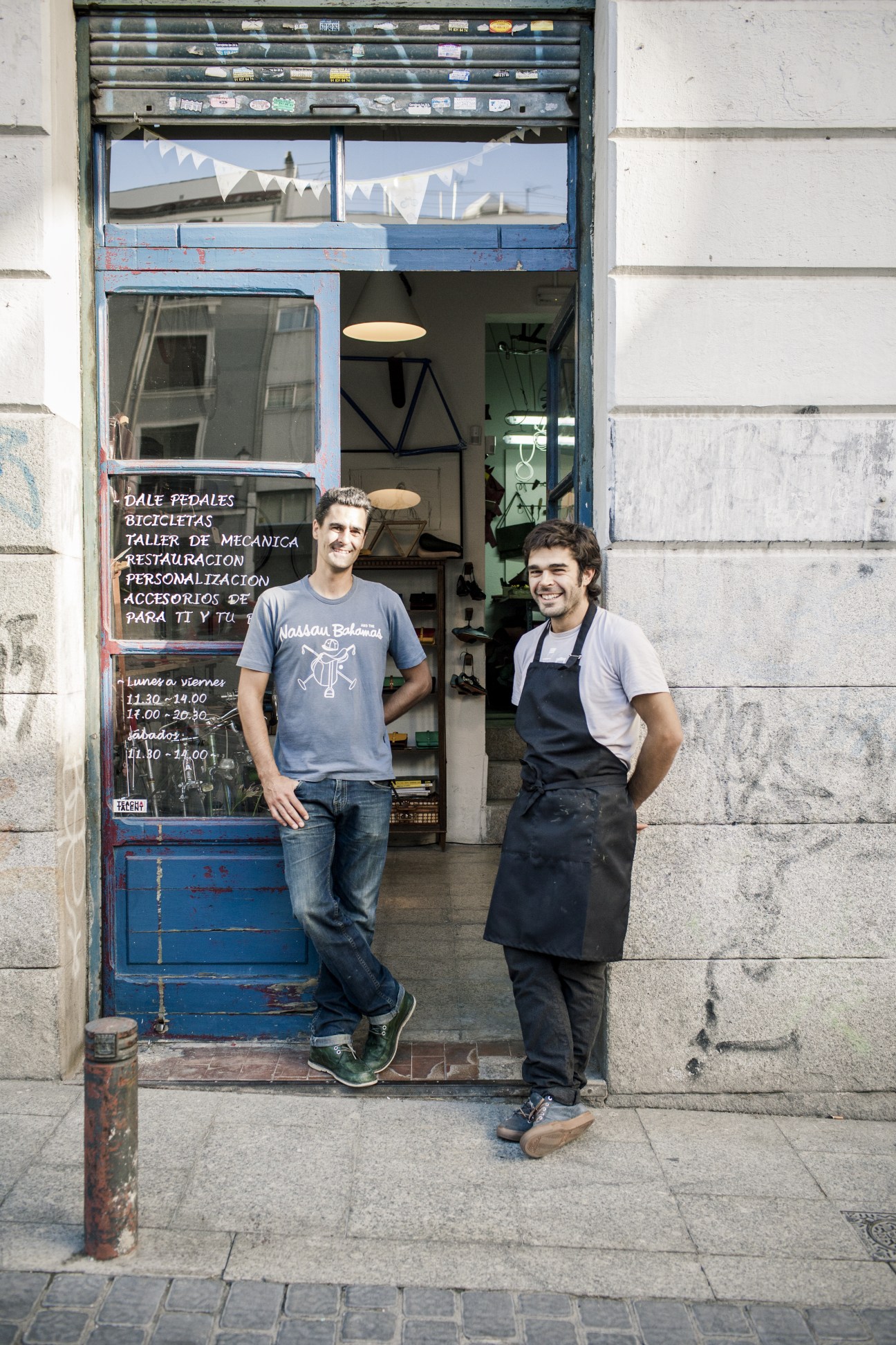 Two men stand in the doorway of Dale Pedales, a Madrid bicycle repair and leather goods shop with a weathered blue storefront.