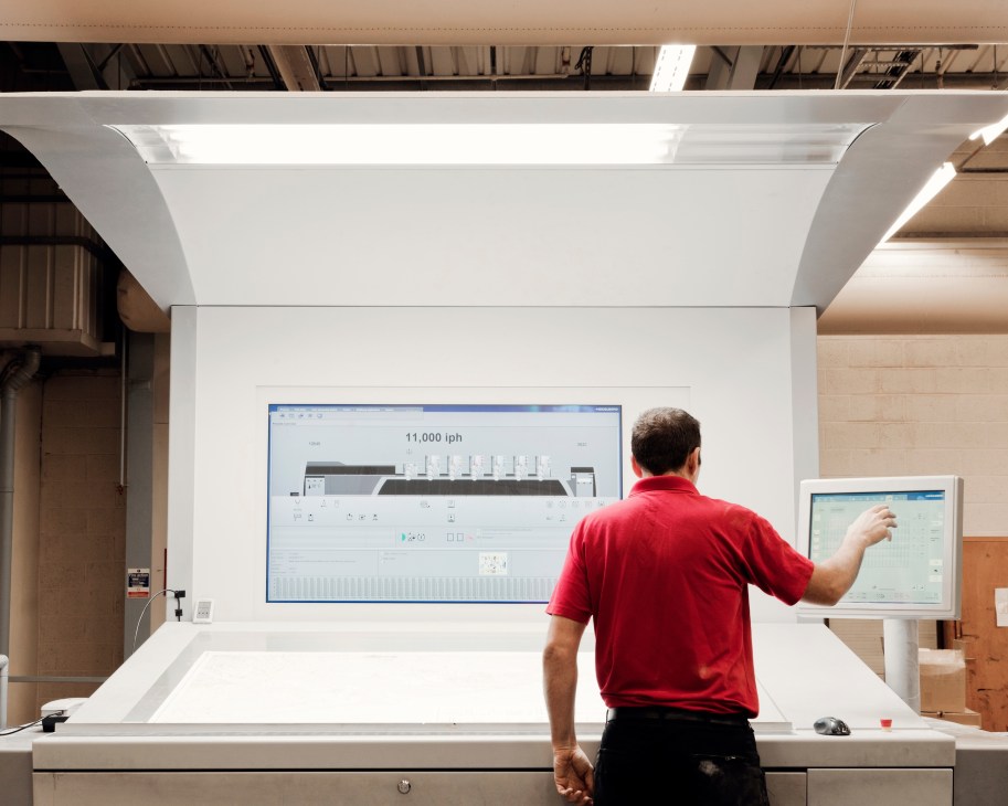 Man in red shirt operating large lithographic printer displaying technical specifications at Butler Tanner & Dennis printing facility.