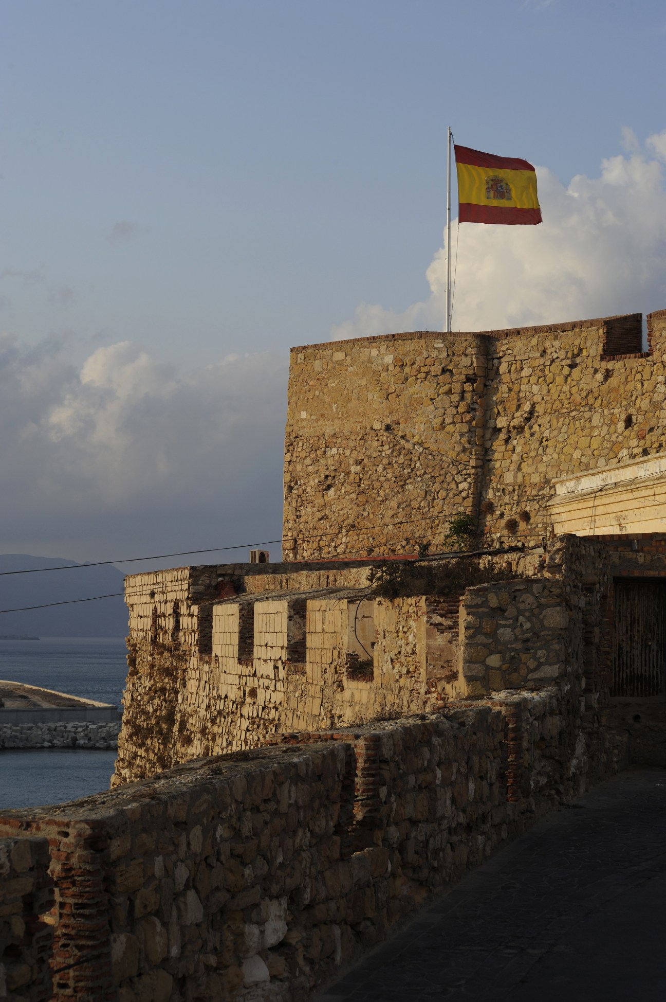 Spanish flag flying above a weathered stone fortress overlooking the Mediterranean Sea in Melilla's old town.