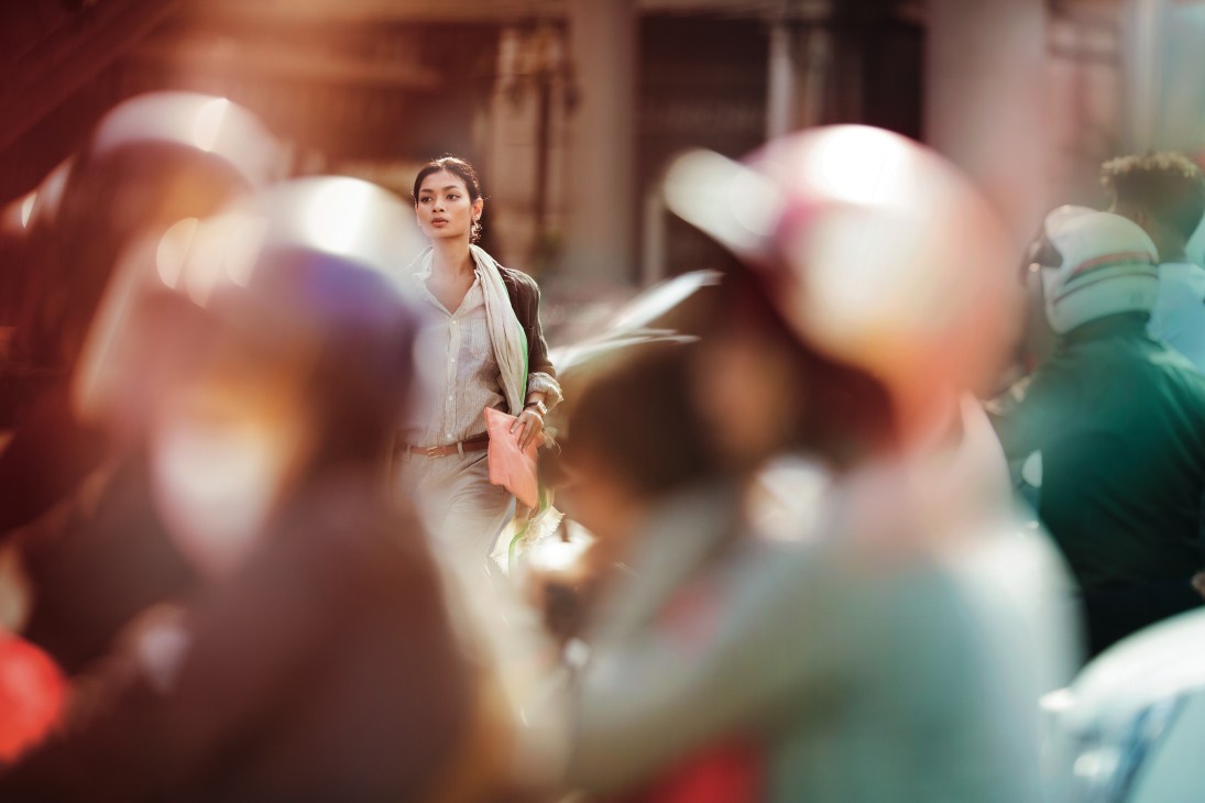 Woman in Toujours jacket and shorts walking through busy Bangkok street surrounded by blurred pedestrians.