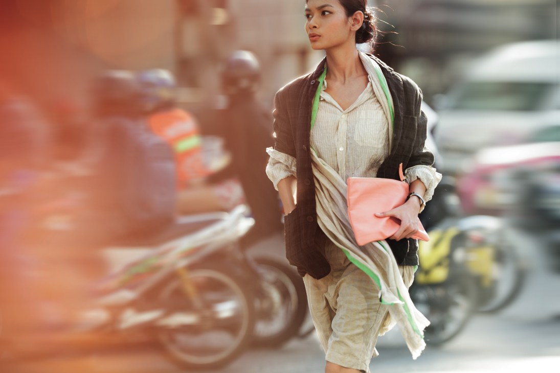 Woman in brown jacket and cream shorts holding coral bag walking past Bangkok taxis