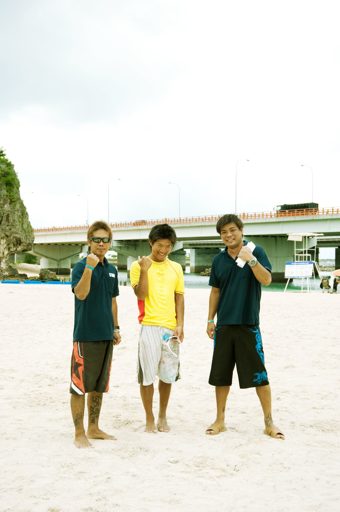Three young male lifeguards posing on sand at Naminoue Beach with a bridge in the background.