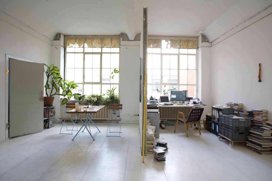 A bright artist studio with high ceilings, large windows, workspace with laptop, plants on windowsills, and stacked books on the floor.