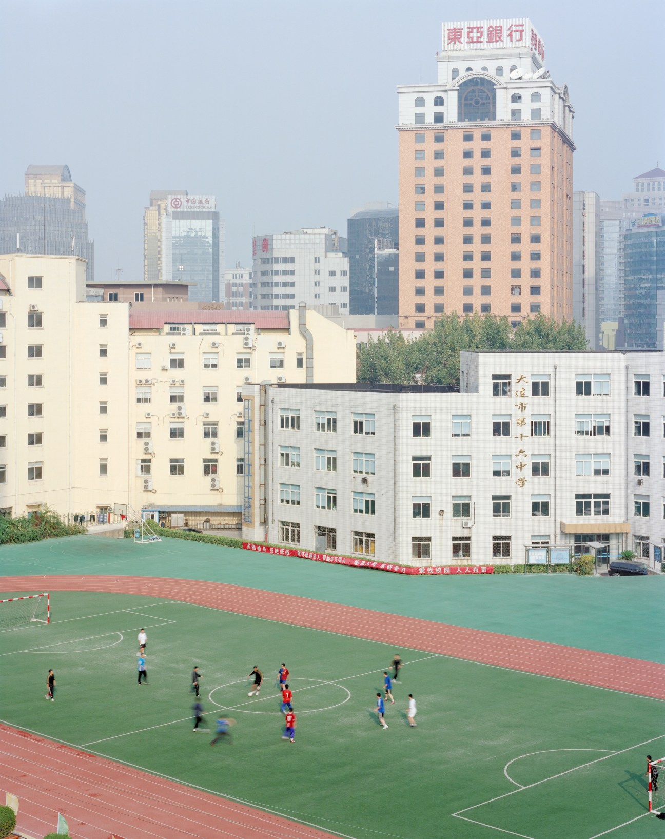 Children playing soccer on a green field with Dalian's modern skyline and colonial-era buildings behind them.