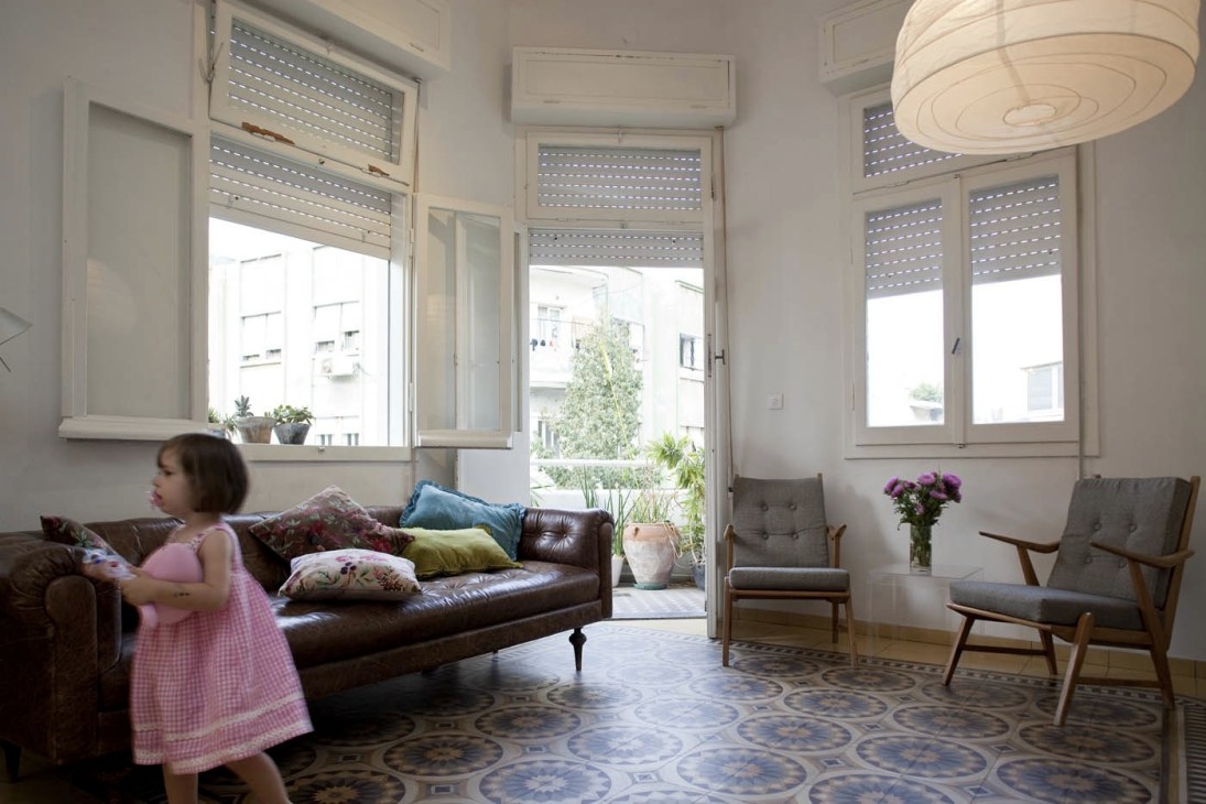 Woman in pink dress sits on brown leather sofa in bright living room with patterned floor and modern furnishings.