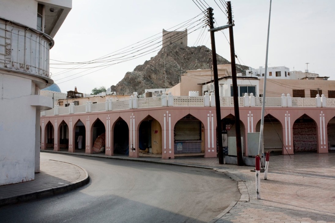 Pink-painted arcade building with arched openings in old Muscat, with white buildings and rocky mountains beyond.