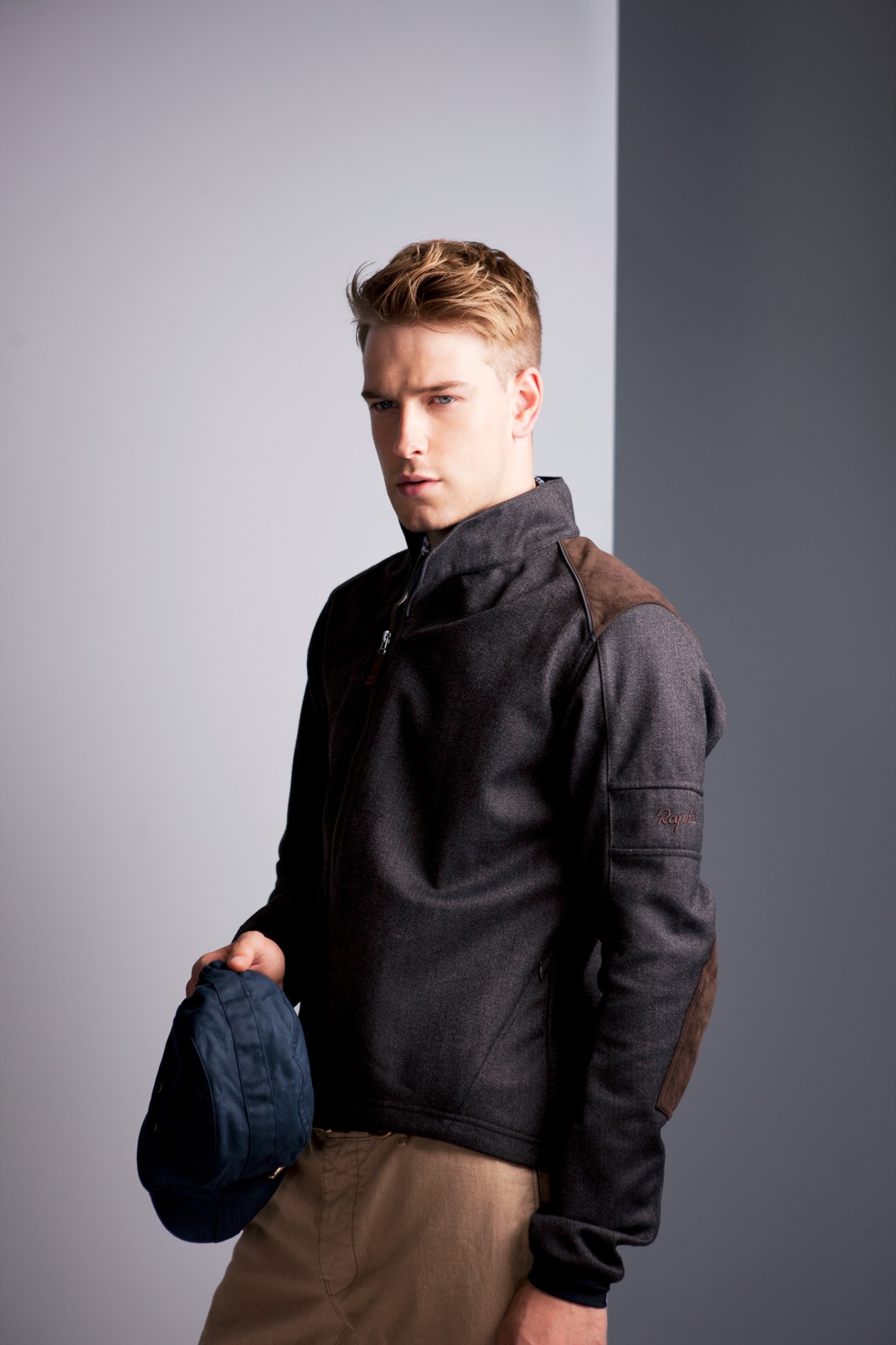 Man wearing Rapha dark tweed cycling jacket with brown shoulder patch and holding blue cap.
