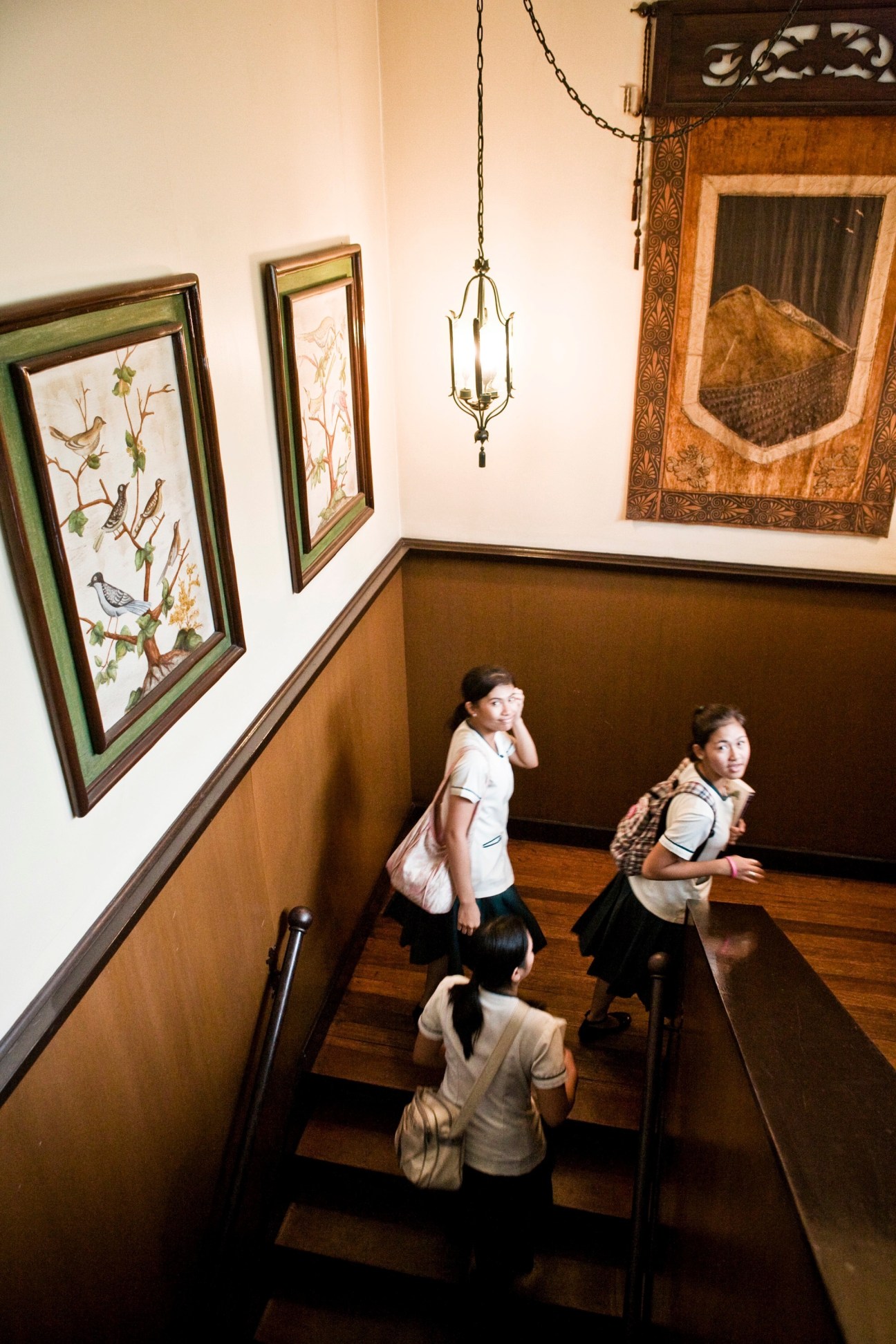Three young women walk down a staircase with ornate artwork and a hanging lantern at Punlaan School.
