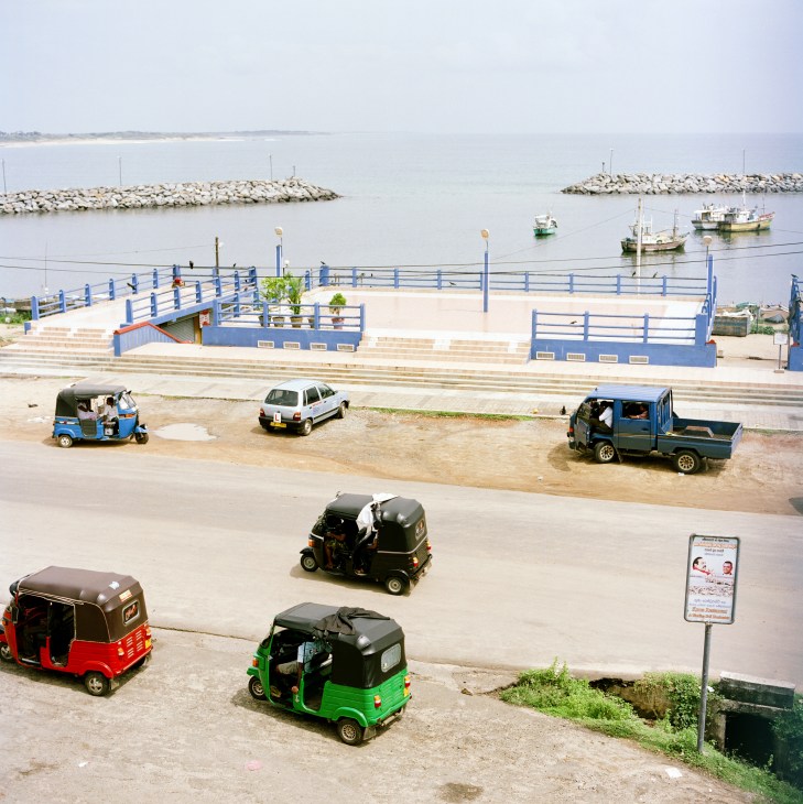 Hambantota seafront with fishing boats, tuk-tuks and vehicles parked near blue-railed waterfront promenade.