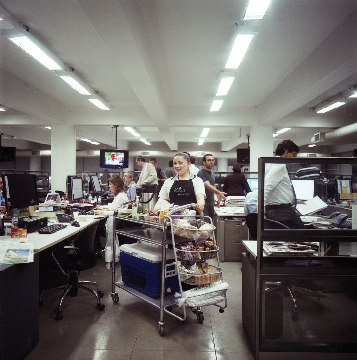 A vendor pushes a cart with snacks and beverages through a busy newsroom filled with desks and journalists.