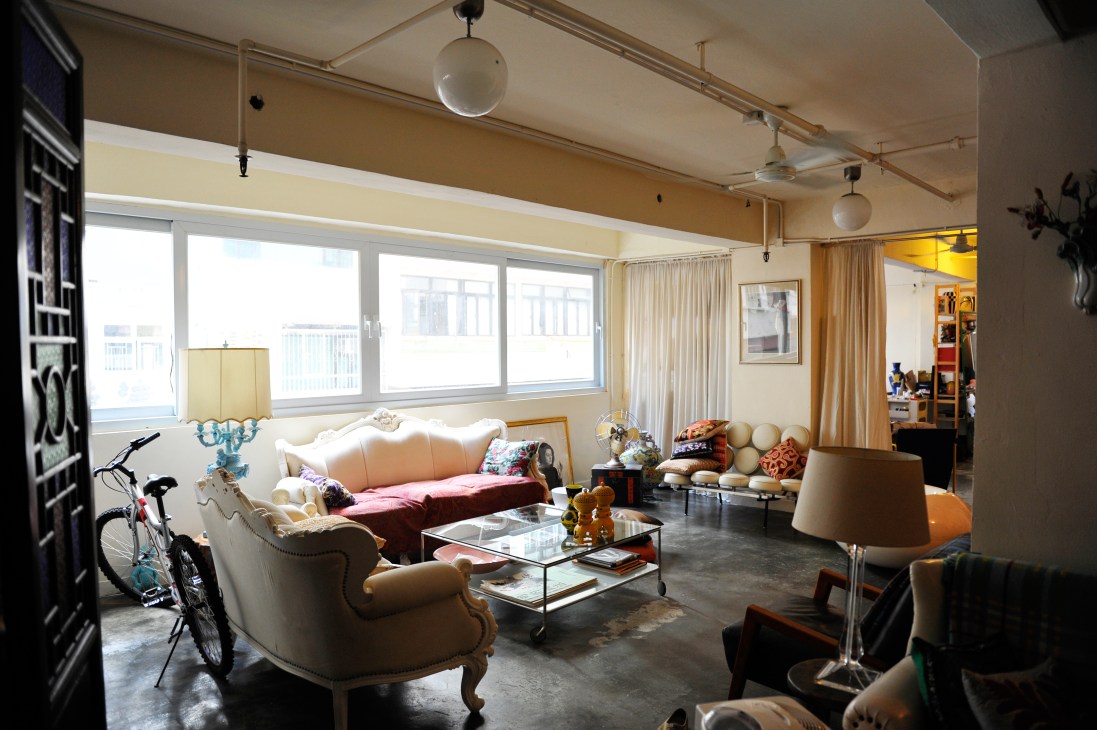Spacious loft-style living room with vintage furniture, large windows, and eclectic decor including a bicycle and colorful cushions.