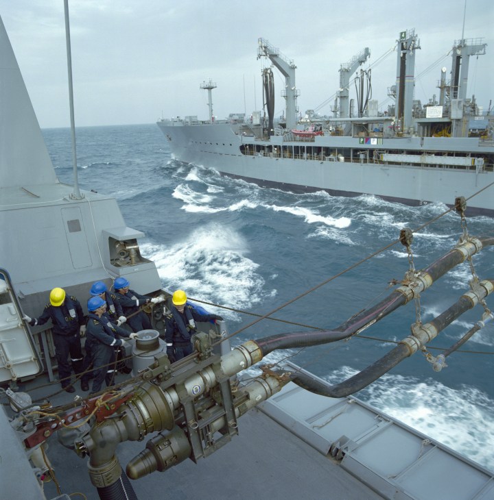 Navy crew members in hard hats coordinate fuel replenishment lines between two ships at sea.