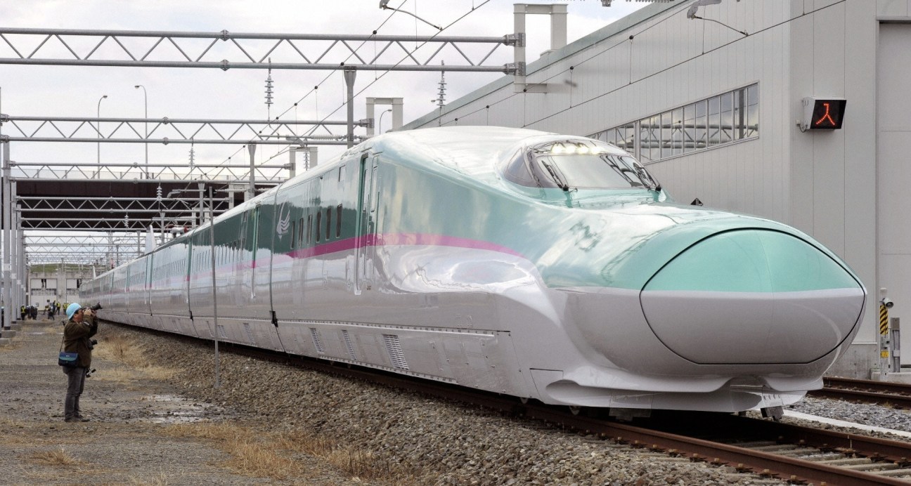 A white and turquoise Shinkansen bullet train at a station platform under overhead electrical lines.