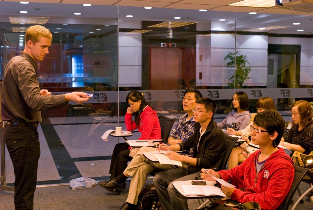 EF English teacher instructs a class of young Chinese students seated in a modern classroom with glass partitions.