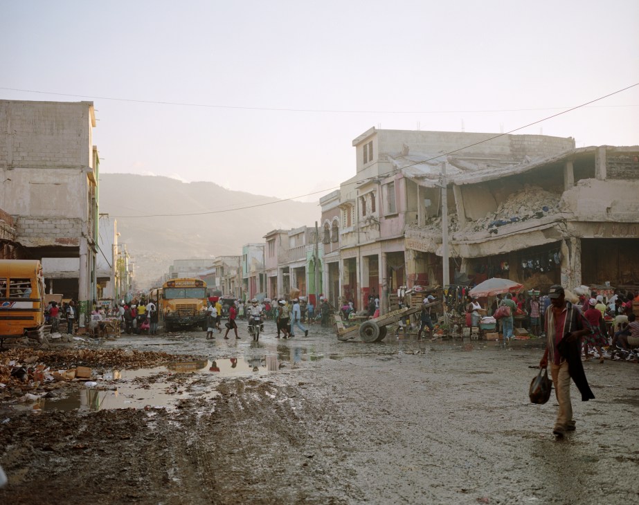 Damaged buildings and rubble line a wet street in downtown Port-au-Prince with residents and vehicles visible.