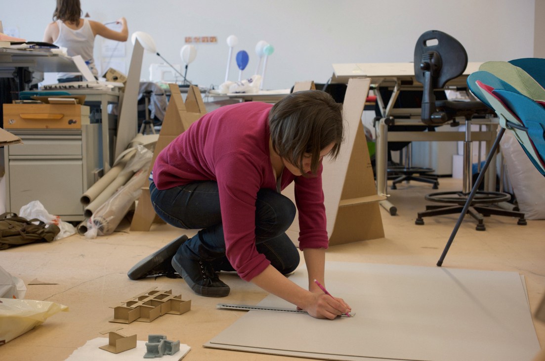 A student in a burgundy shirt crouches while sketching on a large sheet of paper at an ECAL workshop desk surrounded by cardboard models and design materials.
