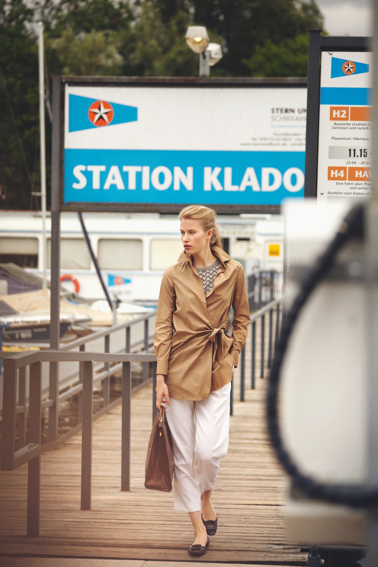 Woman in camel coat and white trousers walking on wooden dock past Station Klado sign at Wannsee lake.