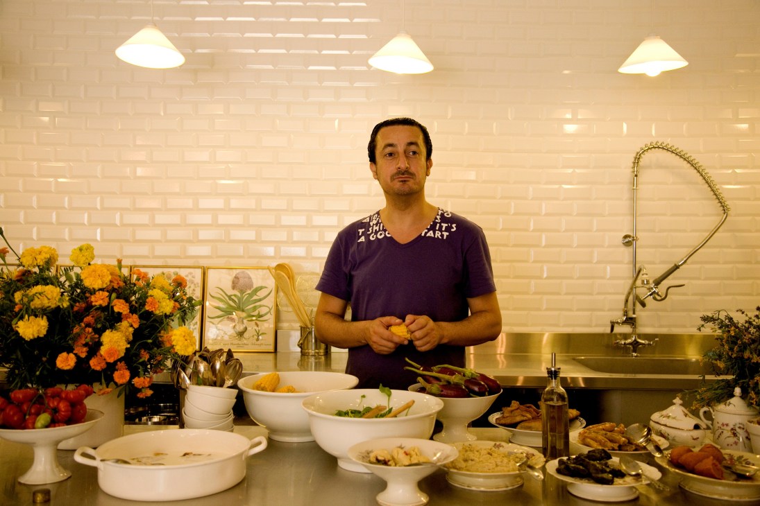 A man in a purple shirt stands in a bright kitchen with fresh vegetables, flowers, and traditional Lebanese dishes displayed on the counter.
