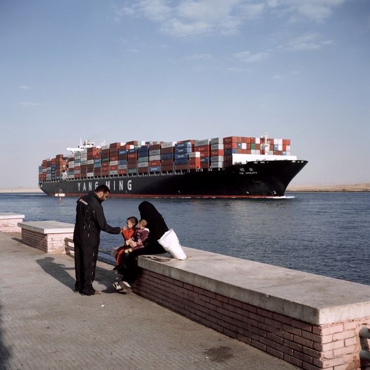 A large container vessel passes through the town of Suez as locals watch from the waterfront.